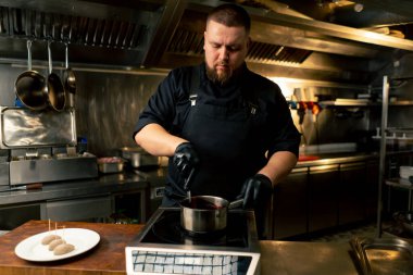 Siyah ceketli profesyonel bir aşçı sosu tencereye karıştırıyor.