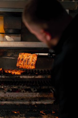 Sıcak ızgara fırınının yanında siyah ceketli profesyonel bir aşçının yakın çekimi kaburgaları devirir.