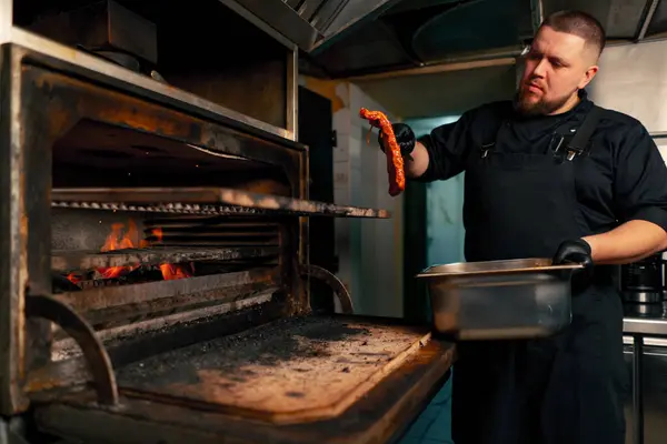 在一个专业的厨房里 穿着黑色夹克的厨师在一个热烤箱附近放着腌制的排骨 — 图库照片