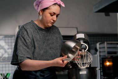 Profesyonel bir mutfakta, demir bir masanın üstüne bir fırıncı bir miksere un döker.