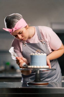 Profesyonel bir mutfakta, mavi pastayı kremayla süsleyen bir kadın fırıncının resmi.