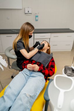 Bir dişçi bir kızın diş muayenehanesinde bir kızın ağız boşluğunu muayene eder.