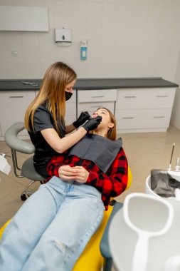 Bir dişçi bir kızın diş muayenehanesinde bir kızın ağız boşluğunu muayene eder.