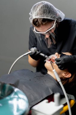 Diş muayenehanesinde şapkalı bir kadın dişçi düzenli olarak genç ve güzel bir kızın dişlerini temizler.