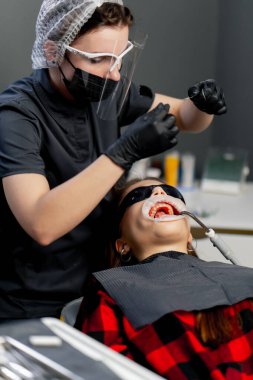 Bir dişçi ofisinde kepli kadın dişçi genç ve güzel bir kıza diş ipi kullanıyor.
