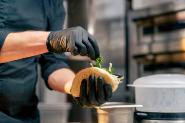 Siyah eldivenli bir şef maşayla çöreğe farklı malzemeler koyar.