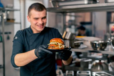 the chef in the kitchen of the establishment examines in his hands a ready-made burger lying on a black plate clipart