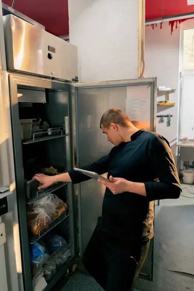 シェフは黒い制服のプロのキッチンに立って 冷蔵庫のリストにあるものを調べる — ストック写真
