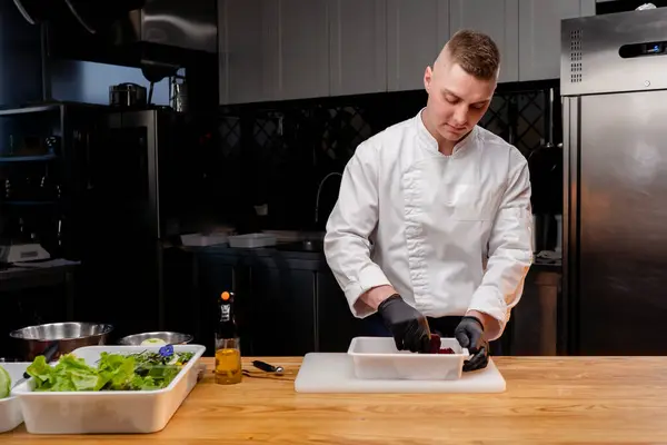 A chef in a white jacket in a professional kitchen mixes beets in a white bowl