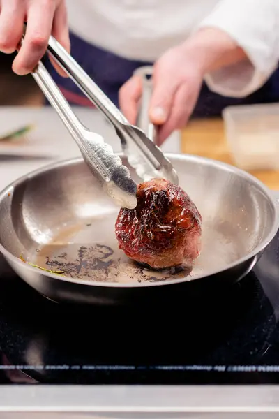 close-up in a professional kitchen with tongs holding a beef steak over a hot frying pan with oil