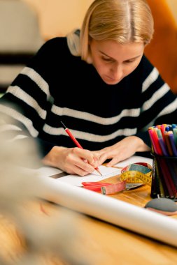 Bir dikiş atölyesinde genç bir kız kırmızı kalemle gelecekteki kıyafetlerin bir taslağını çiziyor.