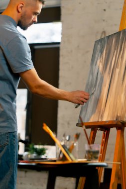 Bir sanat atölyesinde mavi tişörtlü bir sanatçı palet bıçağıyla tuval üzerinde geniş vuruşlar yapar.