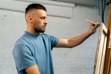 Bir sanat atölyesinde mavi tişörtlü bir sanatçı geniş ve düz bir fırçayla tuval üzerinde geniş vuruşlar yapar.