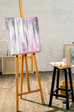 Sanat atölyesinde üzerinde palet ve fırça olan taburesinin yanında üç ayaklı bir tablo var.