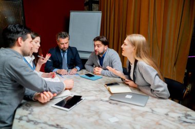 Ofis çalışanları, müdürün önderliğindeki kırmızı duvarlarla bir konferans salonunda oturmuş aktif olarak tartışıyorlar.