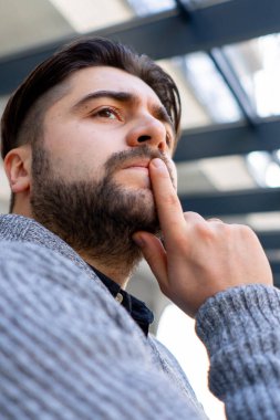Sakallı bir delikanlının yakından çekilmiş fotoğrafı. Düşünceli bir şekilde pencereden bitkilerin arasına bakıyor.