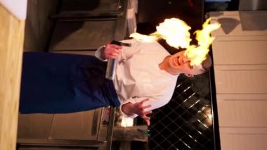 Profesyonel bir mutfakta dikey bir video ceketli bir şef yanan bir ocakla ateşli bir şov yapıyor.
