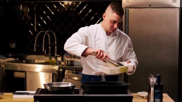 穿着白色制服的厨师在一个专业的厨房里把几块蔬菜放在有钳子的煎锅里 — 图库视频影像