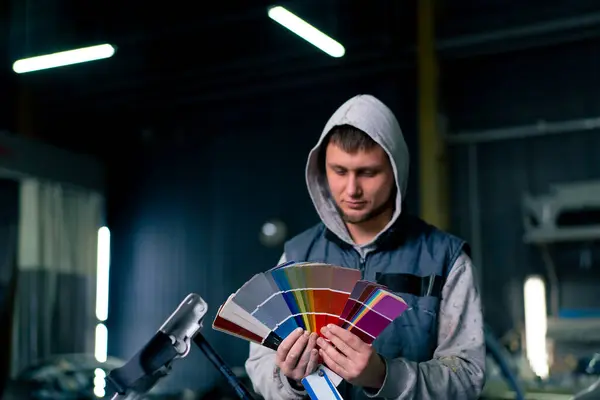 A paint master at a service station selects a paint color on a colored paper palette against the background of his car
