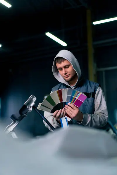 A paint master at a service station selects a paint color on a colored paper palette against the background of his car