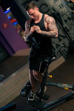 Spor salonunda siyah tişörtlü, dövmeli genç yakışıklı bir adam egzersiz makinesine karın kaslarını pompalıyor.