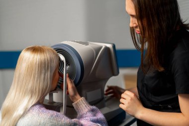 Göz hekimliği kliniğinde bir kadın doktor parmaklarıyla bir kıza göz altındaki deriyi nasıl sıkılaştıracağını gösterir.