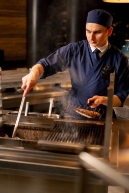 Bir Japon restoranında mavi üniformalı bir şef mutfaktaki ızgarada balık kızartıyor.