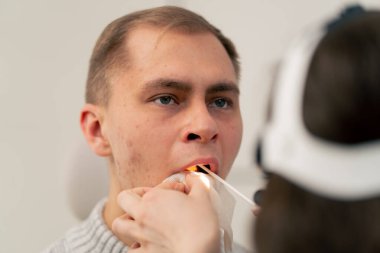 Klinikte genç bir doktor kulağının burun kısmı hastanın boğazını muayene ediyor.