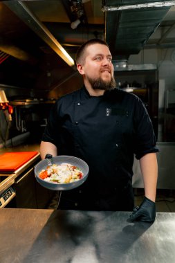 Profesyonel mutfakta şef elinde hazır salatayla kameraya gülümsüyor.