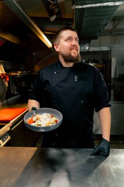 Profesyonel mutfakta şef elinde hazır salatayla kameraya gülümsüyor.