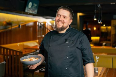 Siyah ceketli bir restoran şefi hazır salatayla gülümsüyor.