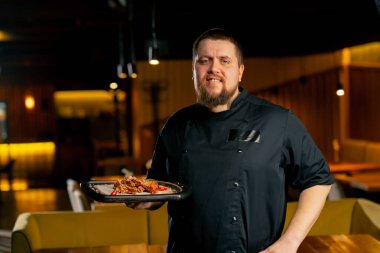 Siyah ceketli bir restoran şefi hazır tavuk kanatlarıyla gülümsüyor.