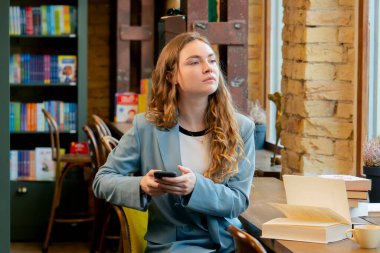Güzel bir öğrenci bir kitapçıda pencerenin kenarında oturur kitap okur ve telefonda sohbet eder.