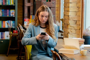 Güzel bir öğrenci bir kitapçıda pencerenin kenarında oturur kitap okur ve telefonda sohbet eder.
