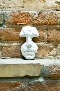 Çıplak tuğla bir duvarın arkasında duran beyaz kil bir maskenin yakın plan görüntüsü.