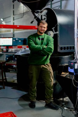 Kahve fabrikasında çalışan bir işçi kızartma makinesinin yanında durup ellerini katlayan kameraya bakıyor.