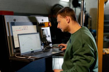 Kahve kızartma fabrikasında yeşil kazaklı bir işçi dizüstü bilgisayarla kızartma makinesini çalıştırıyor.
