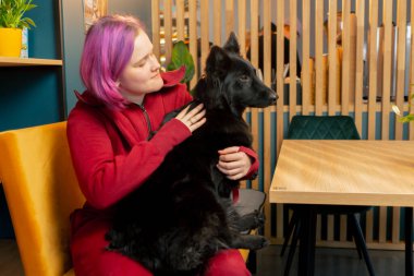 Pembe saçlı genç bir kız bir kafede siyah bir köpeğe sarılıyor.