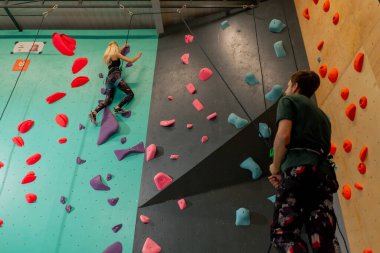 tırmanma duvarında genç bir kız ilk kez duvara tırmanıyor, koç emniyet halatıyla bağlanıyor.