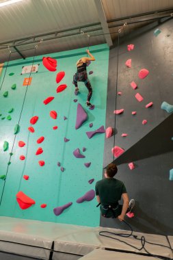 tırmanma duvarında genç bir kız ilk kez duvara tırmanıyor, koç emniyet halatıyla bağlanıyor.