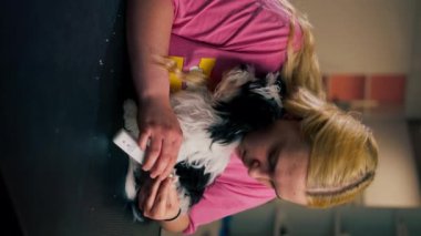 Manikür salonundaki dikey videoda küçük siyah-beyaz bir köpek, tırnak törpüleme prosedürünün yanında.