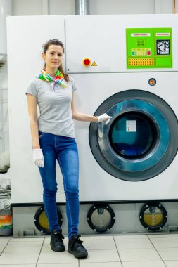 Profesyonel kuru temizlemeci genç kız kameraya bakıyor ve profesyonel bir çamaşır makinesinin yanında poz veriyor.