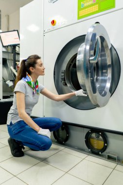 Profesyonel kuru temizlemeci genç kız kameraya bakıyor ve profesyonel bir çamaşır makinesinin yanında poz veriyor.