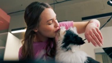 Bir kuaförde pembe tişörtlü genç bir kız siyah-beyaz bir köpeği öpüyor.