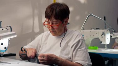 Bir makinenin dikiş atölyesinde yaşlı terzi kadın siyah bir kumaşı kesiyor.