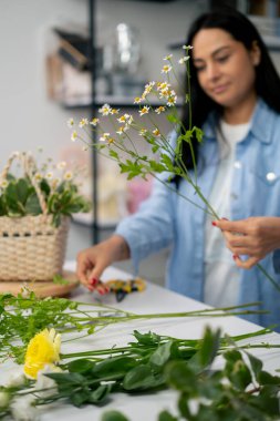 Bir çiçekçide, beyaz bir masanın yanında bir kız çiçek aranjmanı topluyor sepette bir buket çiçek.