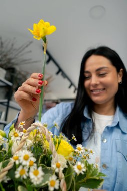 Bir çiçekçide beyaz bir masanın yanındaki kız sarı nergislerden bir çiçek aranjmanı topluyor.