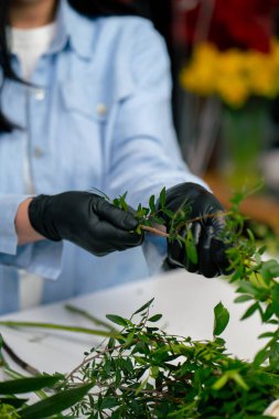 Bir çiçekçide, beyaz bir masanın yanında bir kız bir sepet çiçeği süslemek için zeytin ağacının dalını buduyor.