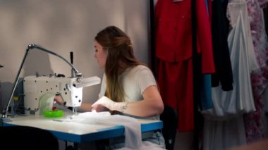 Dikiş atölyesinde genç bir kız dikiş makinesi ve beyaz kumaşla çalışıyor.