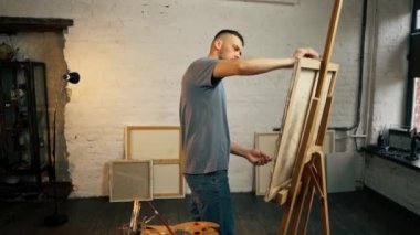 Bir sanat atölyesinde mavi tişörtlü bir sanatçı etkin bir şekilde fırçayla tuvale resim yapıyor.